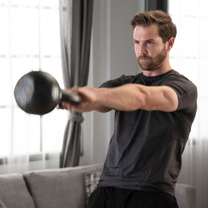 Lifestyle shot of a man performing a Hammertone kettlebell swing in a home living room setting. He wears a grey t-shirt and black shorts, swinging a black kettlebell upwards with both hands. The kettlebell is blurred to indicate the speed of the motion.