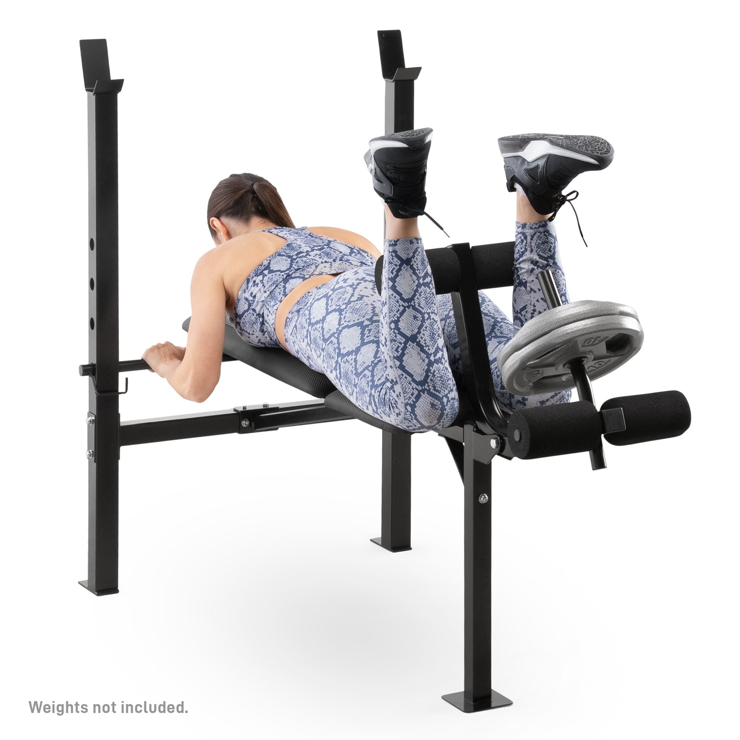 Studio shot of a woman performing a lying leg curl on the Marcy Standard Weight Bench MWB-6501 weight bench. She lies face down on the black vinyl padding, hooking her ankles under the top foam rollers of the leg developer to lift the loaded weight plates. A disclaimer at the bottom reads, "Weights not included.".