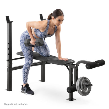 Studio shot of a woman performing a single-arm dumbbell row on a Marcy Standard Weight Bench MWB-6501. She kneels on the bench padding with one leg while lifting a hexagonal dumbbell with one hand. The bench features a leg developer attachment loaded with weight plates at the front and vertical barbell uprights at the rear. A disclaimer at the bottom reads, "Weights not included.".